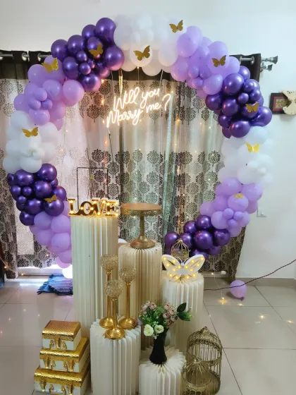 This is one of my signature proposal setups, designed to create a magical moment. It features a 'Will you Marry me?' neon sign set against a full circular balloon arch in shades of purple and white, with elegant white pedestals ready for displaying rings, flowers, or gifts.