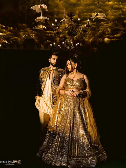 A dramatic, low-key portrait under a stunning chandelier. The dark background makes their golden attire shine, creating a moody and opulent feel.
