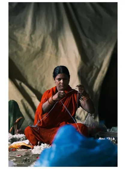 6:53am: A female artisan sits against a canvas backdrop, her hands expertly weaving garlands. The dramatic lighting highlights her focused expression and the delicate nature of her craft.