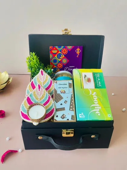 A stylish and affordable hamper in a black suitcase box. This gift set includes a leaf-shaped beaded T-light holder, green tea, a chocolate bar, and snacks, perfect for corporate gifting under ₹1000.