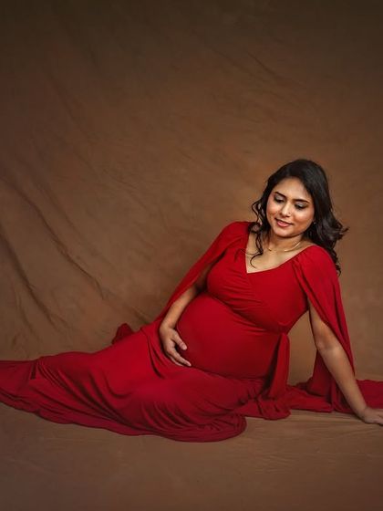A peaceful, full-length portrait of the mother-to-be seated on the floor. The flowing red gown pools around her, creating a beautiful and serene image against a simple brown backdrop.