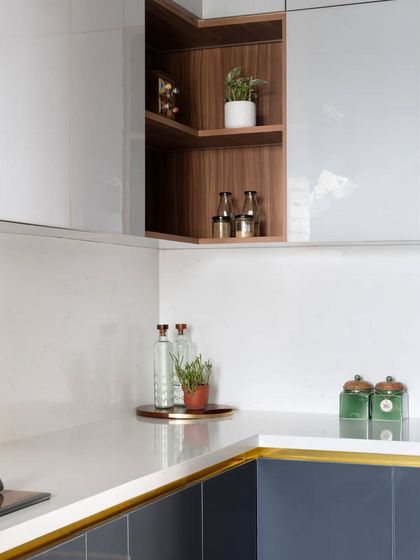 In this kitchen, a familiar fusion of white, grey, and blue exudes a modern yet classic charm. An open corner shelf in a wood finish breaks the monotony of the glossy cabinets.