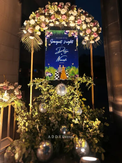 A stylish welcome sign for Divya & Nimish's Sangeet Night, set within a lush arrangement of greenery and disco balls, topped with a floral arch.