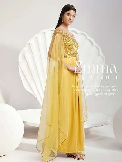 The Emma Jumpsuit in a vibrant yellow. This unique piece features an embellished off-shoulder bodice, dramatic cape sleeves, and wide-leg pleated trousers for a modern and graceful statement.