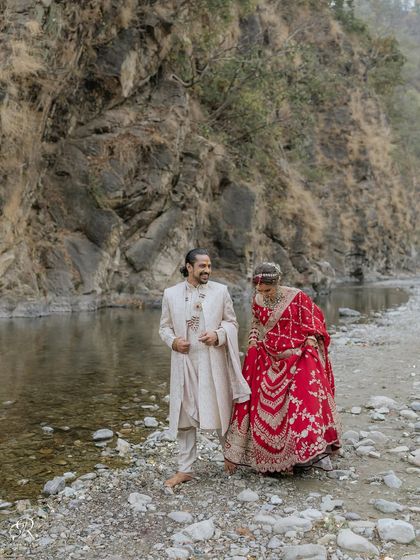 A candid shot of the couple walking along the rocky riverbank after their wedding. Her beautiful red lehenga stands out against the natural scenery, and their laughter is infectious.