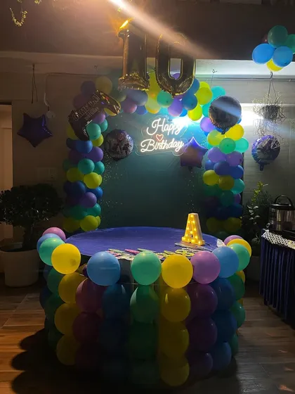 The cake table for the rooftop party, surrounded by a colorful balloon arch and a 'Happy Birthday' neon sign. The number '10' foil balloon marks the special milestone.