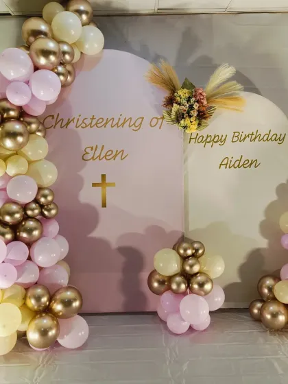 A combined Christening and birthday celebration. The elegant pink backdrop is decorated with a pastel pink and gold balloon garland, with names and a cross decal.