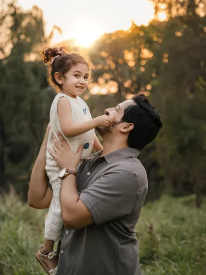 A father holds his daughter up, and she playfully touches his face. This image captures a sweet, interactive moment during their family photoshoot.