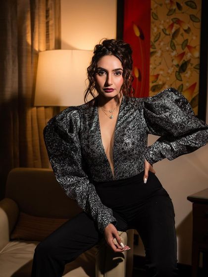An indoor portrait of Ragini Dwivedi in a stylish silver and black top. The warm, soft lighting from the lamp creates an intimate and sophisticated mood.