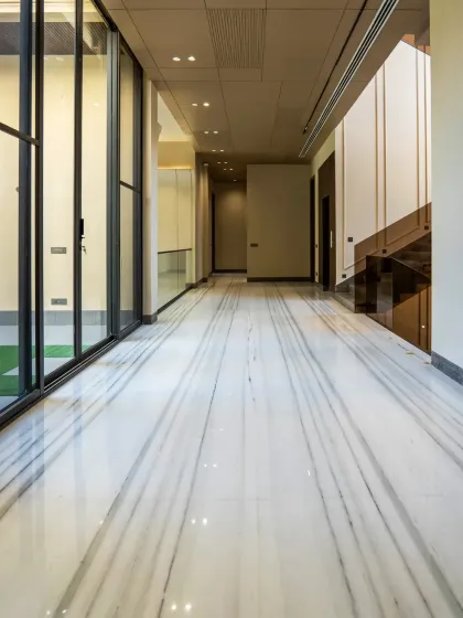 The long corridor in this farmhouse is lined with large glass panels on one side, overlooking an internal courtyard. The striped white marble floor enhances the sense of length and perspective.