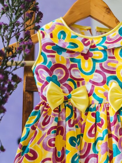 A vibrant, abstract-print dress in shades of yellow, pink, and teal. The double bows at the waist add a sweet, symmetrical detail to this fun and energetic design.