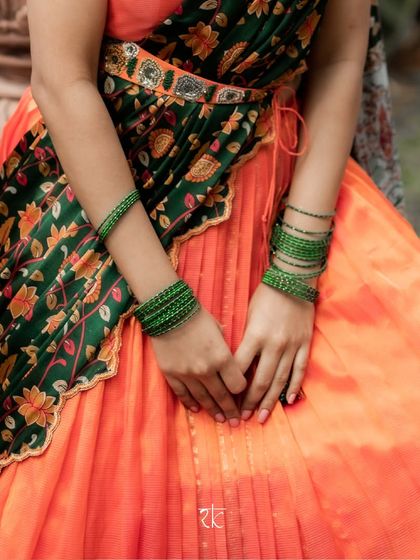 The pleated silk skirt is cinched with an embroidered belt, and the contrasting green dupatta with Kalamkari prints creates a striking look.