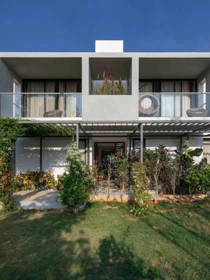 The front facade of the SAMAM House, designed to be a welcoming space for a family and their pets. The design balances private upper-level balconies with a green, accessible front yard, shaded by a modern pergola structure.
