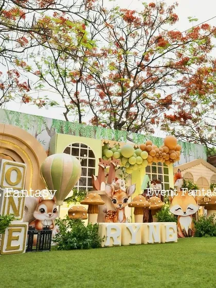 An enchanting woodland first birthday for Ryne, set in a beautiful green lawn. This wide shot captures the full magical scene, with large cutouts of a squirrel and deer, a charming woodland house backdrop, and whimsical mushroom props surrounded by lush trees.