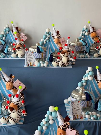 For a child's first birthday, I designed these adorable jungle-circus themed party favors. Each setting includes a plush animal wearing a party hat, a jar of treats, and a balloon-like arrangement in blue and white.