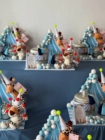 For a child's first birthday, I designed these adorable jungle-circus themed party favors. Each setting includes a plush animal wearing a party hat, a jar of treats, and a balloon-like arrangement in blue and white.
