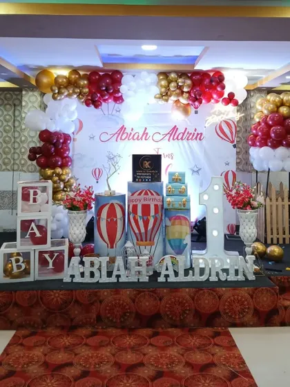 A whimsical hot air balloon theme for a first birthday. The stage includes a custom printed backdrop, balloon sculptures shaped like hot air balloons, and a large light-up number "1".