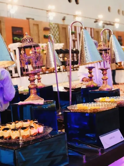 An elaborate dessert counter featuring a variety of Indian sweets. The blue and gold theme with decorative lamps creates a rich and inviting display for guests with a sweet tooth.