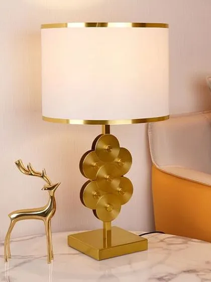 This elegant lamp features a unique base made of stacked golden discs on a square pedestal. Paired with a white and gold-trimmed drum shade, it adds a touch of modern luxury and brightens up any space.