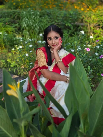 The vibrant red and white of the Bengali saree pop against the lush greenery. This pose, with her hand gently touching her face, adds a touch of classic elegance to the portrait.