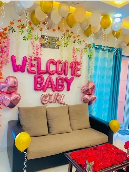 A 'Welcome Baby Girl' decoration in a living room. The setup features pink foil letters, hanging floral vines, and heart-shaped balloons.