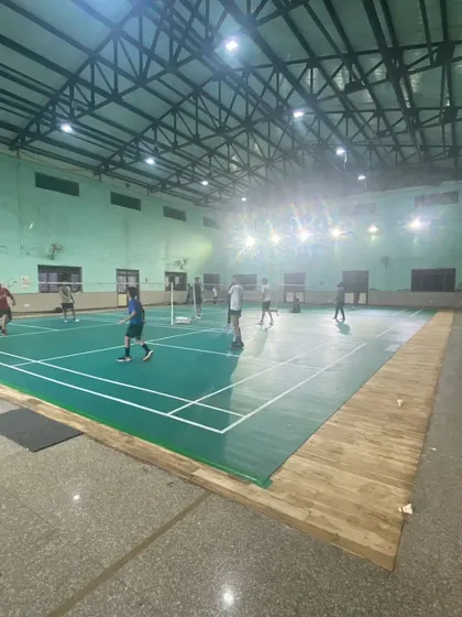 This is our indoor badminton facility at Pitampura, featuring multiple green synthetic mat courts. The space is well-lit for evening and late-night games, with ample room between courts for players.