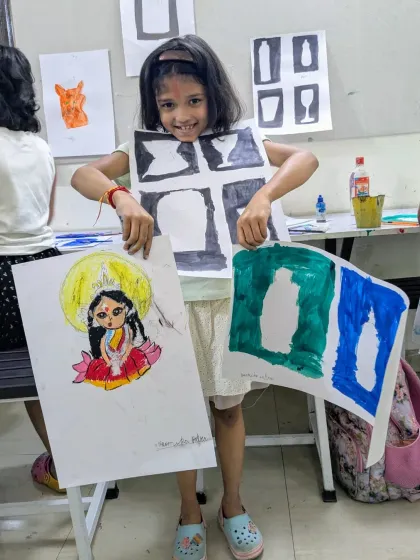 A junior batch student showcasing her diverse work. She holds a colorful painting of the goddess Durga alongside her exercises in negative space, demonstrating a range of skills.