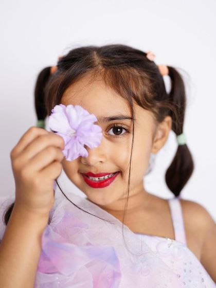 A close-up portrait of a pretty girl with pigtails and red lipstick, playfully holding a flower over one eye.