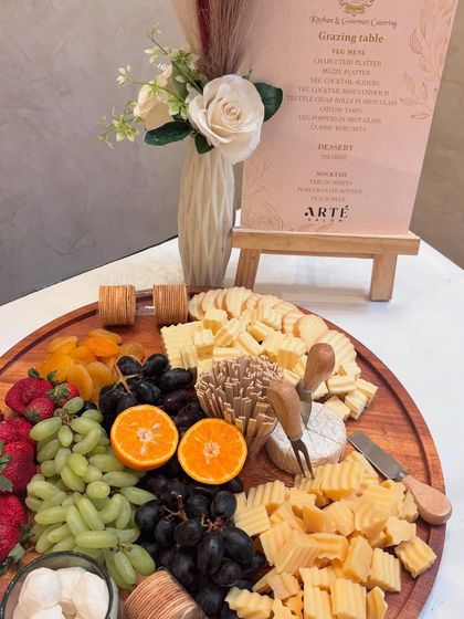 A classic charcuterie board featuring a wheel of brie, assorted hard cheeses, fresh grapes, strawberries, and dried apricots. The accompanying menu details the gourmet selections for the Arte Salon event.