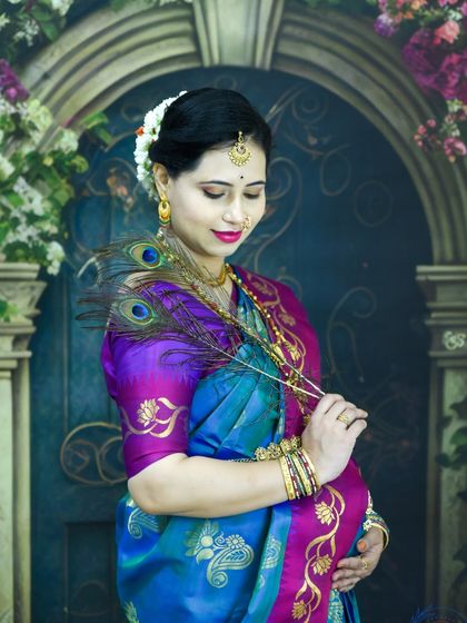 A stunning traditional portrait of a mom-to-be in a silk saree, holding peacock feathers against our floral studio backdrop.