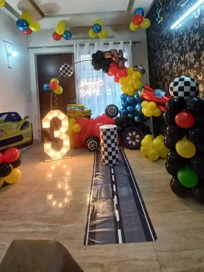A Hot Wheels race car theme for a 3rd birthday. The setup includes a 'race track' floor runner, car-themed cutouts, a marquee number, and a balloon arch in red, yellow, and black.