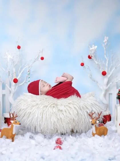 A whimsical Christmas nest. The baby is swaddled in red and placed in a fluffy white nest, surrounded by reindeer and other festive figures for a playful holiday portrait.