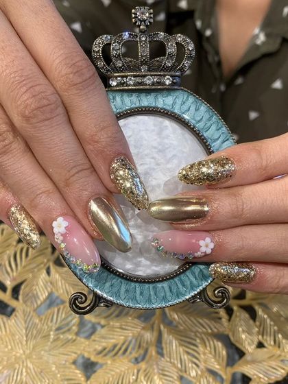 A bold and glamorous set combining gold chrome, gold glitter, and delicate 3D white flowers on long almond-shaped nails.