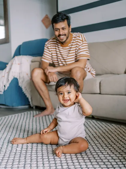 A nine-month-old baby sitting up on the floor, with his dad smiling in the background. A perfect slice of everyday life.
