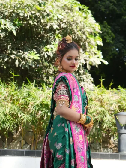 The bride in a natural outdoor setting, her vibrant saree and glowing makeup standing out against the green background.