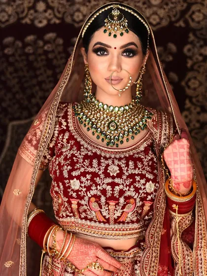 A beautiful bride wearing our rental red velvet lehenga with peacock embroidery. The look is accessorized with a striking green-beaded Kundan necklace and matching maang tikka.