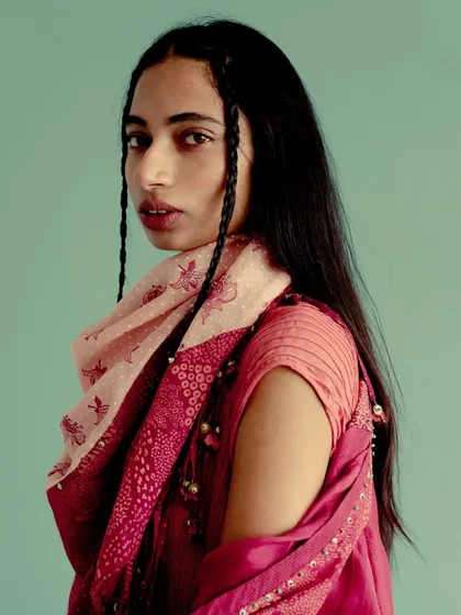 A portrait for Swatti Kapoor against a solid teal background. The warm tones of the pink and red outfit pop against the cool backdrop, highlighting the model's braided hair.