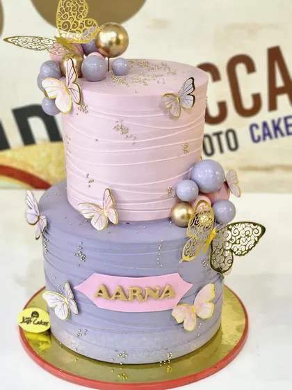 This elegant two-tiered, 4kg birthday cake features a beautiful blend of lavender and pink buttercream. It is adorned with delicate paper butterflies and shimmering gold and lavender spheres, creating a dreamy and sophisticated look for any special occasion.