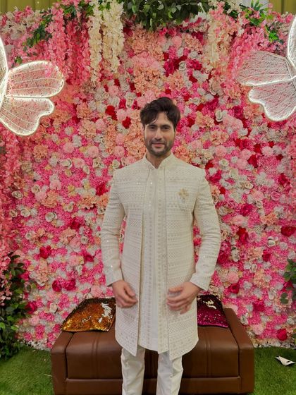 Actor in a stunning off-white Indo-Western sherwani against a floral backdrop. The intricate texture and brooch detail make this a standout piece for any celebration.