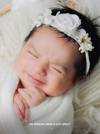 A sweet, scrunched-up smile from a newborn wrapped in white. The floral headband adds a delicate touch to this purely joyful moment.