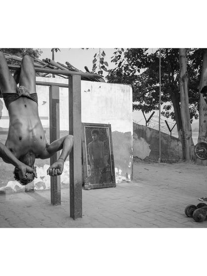 A black and white image of a young man performing an impressive gymnastic move on a pull-up bar at an outdoor gym. Another man lifts weights in the background, showing the community aspect of this space.