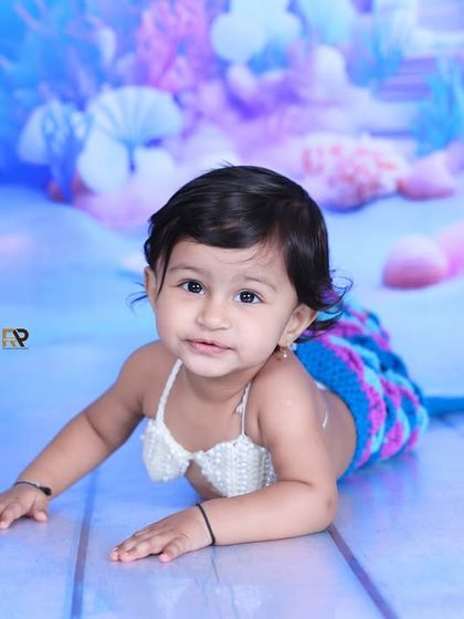 A sweet baby girl lies on her tummy in a mermaid costume, smiling for the camera. The vibrant, colorful underwater backdrop makes this a truly magical portrait.