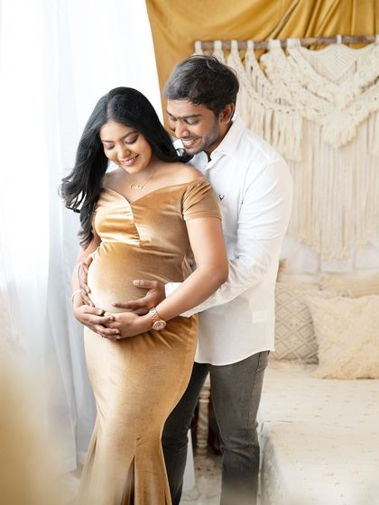 A tender moment between a couple in a bright, boho-themed room. The partner embraces the mom-to-be from behind as they both look down at her baby bump.