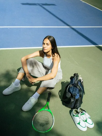 A lifestyle campaign for Lino Perros on a tennis court. This shot creates an aspirational 'athleisure' narrative, showcasing the bag and shoes as part of a stylish, active lifestyle.