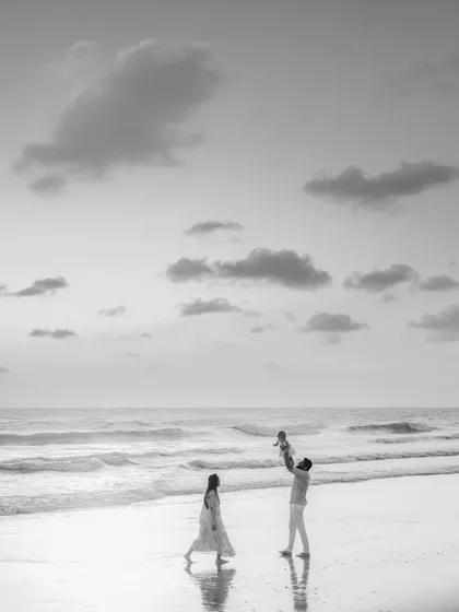 A striking black and white photo of a family on the beach. The father joyfully lifts their child into the air while the mother walks nearby, creating a dynamic and artistic family portrait.
