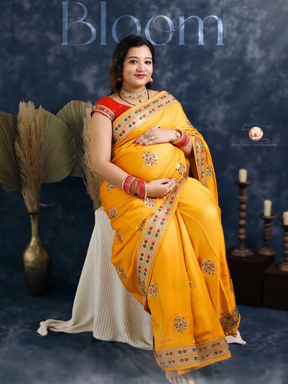 A full-length seated portrait of the mother-to-be in her yellow saree. The word 'Bloom' is overlaid, perfectly describing the radiant phase of motherhood she is in.