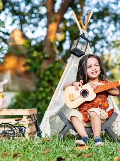 A little boy strums a toy guitar during his outdoor second birthday shoot, featuring a teepee and travel-themed props.