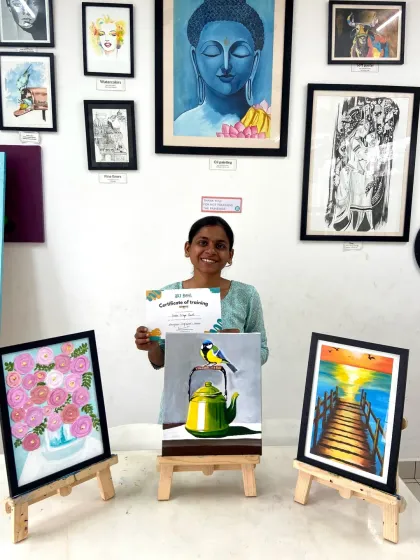 A proud student with her certificate and three acrylic paintings, including a beautiful bird on a kettle and a sunset landscape.