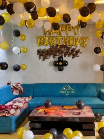 A simple living room setup with black, gold, and white balloons and a "Happy Birthday" foil banner.