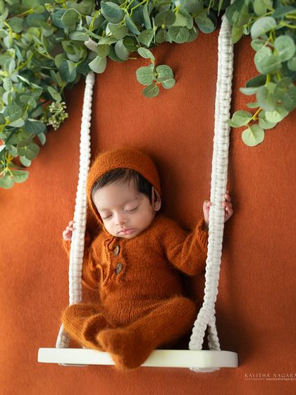 A two-week-old baby rests on a tiny swing, surrounded by greenery. The warm, earthy tones of the outfit and background create a beautiful, natural-light portrait.